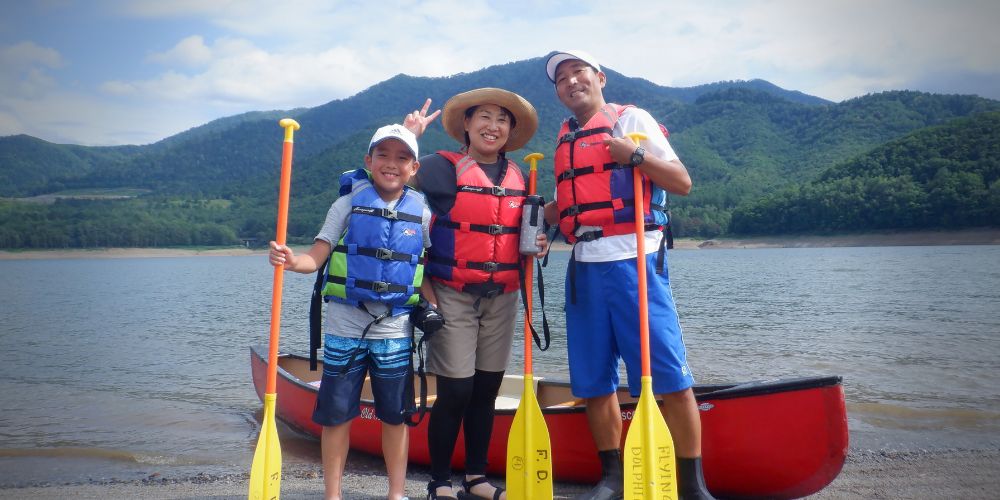 A family of three smiling and posing for a photo with their canoe and paddles on the lake shore.