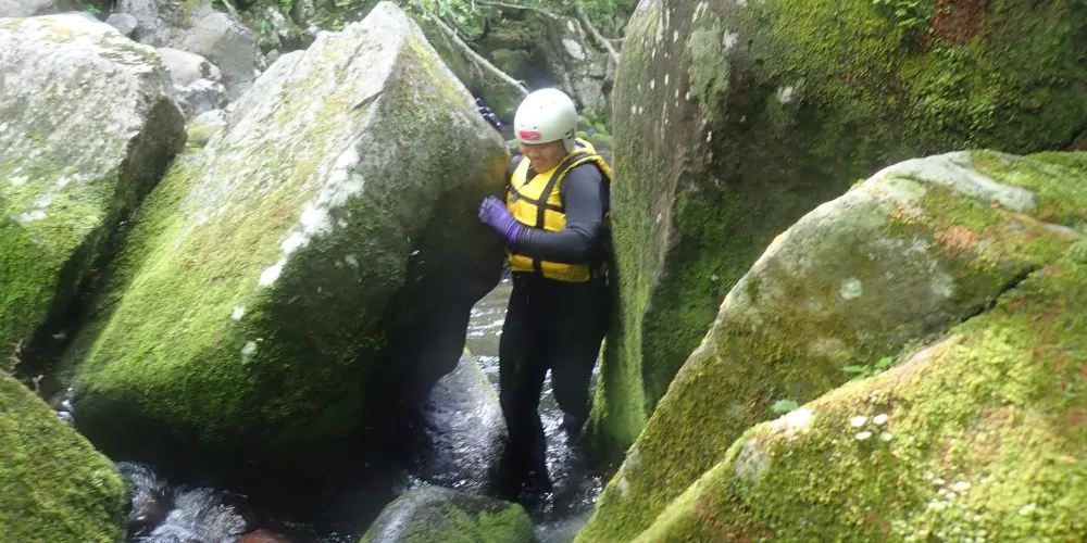 A participant carefully navigating through a narrow passage between large, moss-covered boulders in the wild canyon.