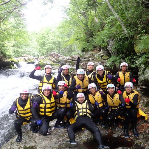 A large team of smiling canyoning participants posing for a group photo by a waterfall.