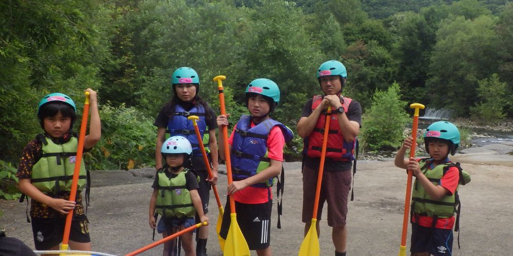 Young participants holding paddles and listening attentively during the pre-tour safety briefing by the river.