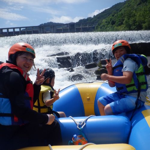 A mother and two children making peace signs on a raft, with a large dam and cascading water in the background.