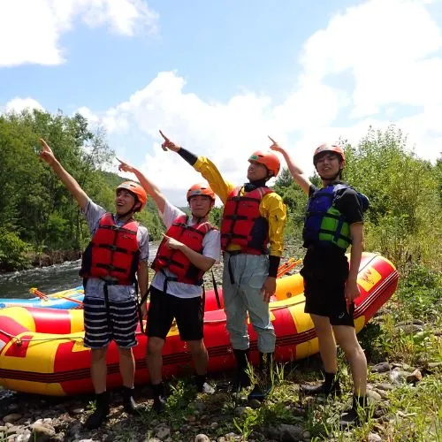 A group of four young rafters pointing up at the sky with excitement on the riverbank, with their colorful raft nearby.