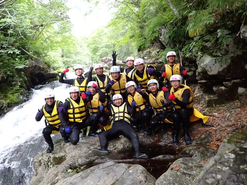 A large, happy group of canyoning participants posing together with thumbs-up and peace signs by a waterfall, celebrating their successful adventure.