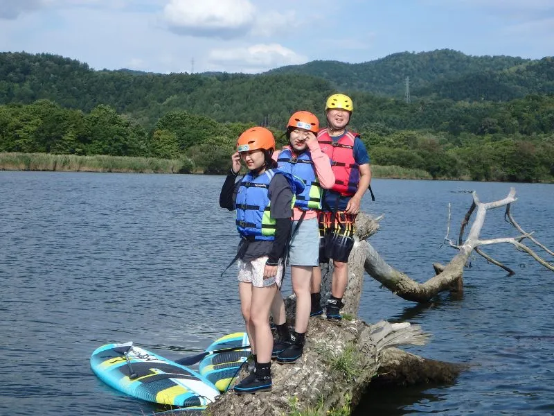 Three happy participants balancing on a large fallen tree in the lake during their SUP tour, highlighting the fun and adventurous aspects of exploring.