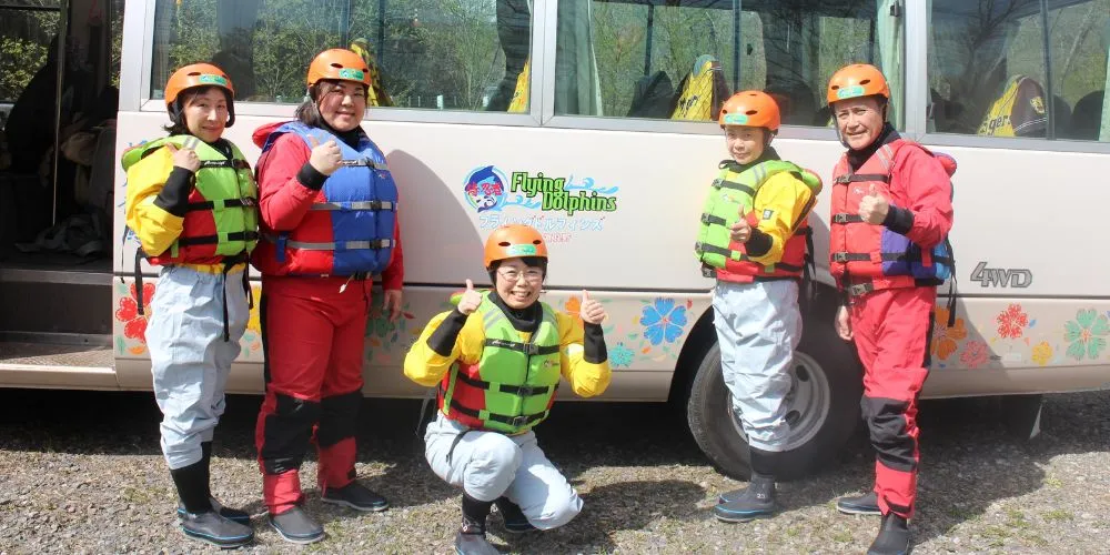 A group of five smiling participants in full rafting gear giving a thumbs-up in front of the transfer bus.