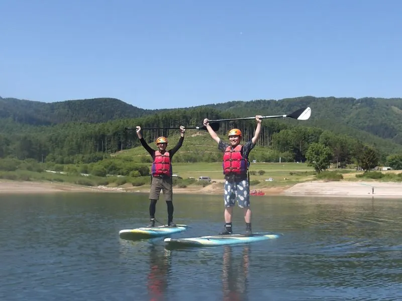 Two participants standing on their paddleboards and raising their paddles in celebration, with a panoramic view of Lake Kanayama behind them.