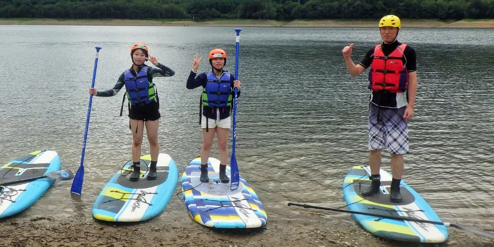 A group of three participants posing with their boards and paddles on the shore before the tour begins.