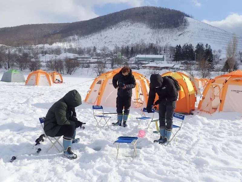 Three people fishing for wakasagi through holes in the ice in front of bright orange tents on a snowy day.