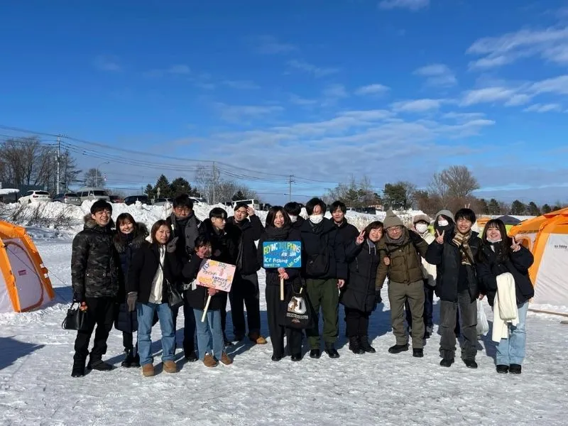 A large group of happy participants posing for a photo on the frozen lake during a Wakasagi ice fishing tour.